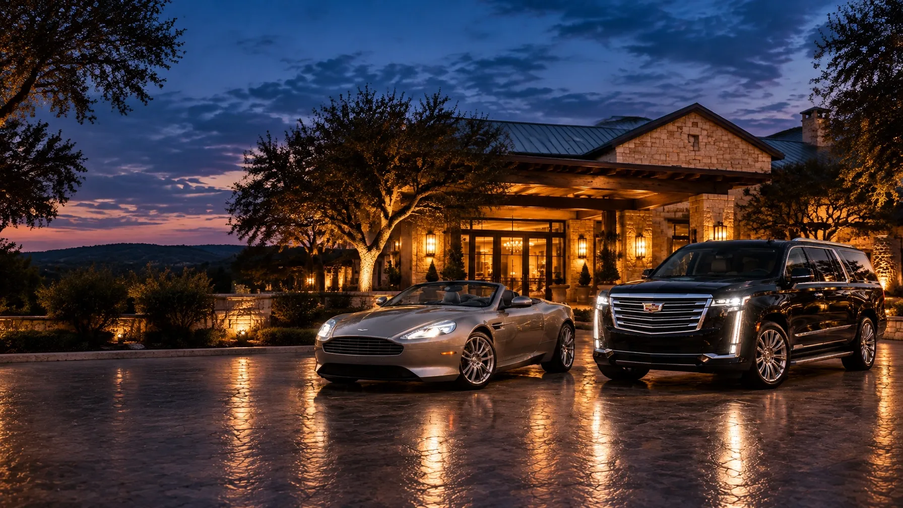 Silver-grey Aston Martin DB9 Volante and black Cadillac Escalade outside a Hill Country hotel at dusk